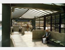 Eingangsbereich der U-Bahn-Station „Rathaus Spandau“ mit einem Glasdach und seitlichen Glaswänden. Eine Rolltreppe führt hinunter, während Passanten und ein Mann auf einer Sitzbank sichtbar sind.