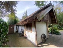 Kleines Fachwerkgebäude mit weiß gestrichenen Wänden und einem Reetdach in einem grünen, bewaldeten Garten. Neben der Tür steht ein Holzrad, und vor dem Haus befinden sich ein Stuhl sowie ein gepflasterter Boden.