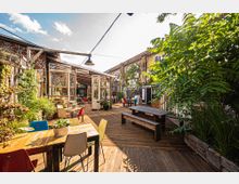 Offener Innenhof mit Holzterrasse, umgeben von Backsteinwänden mit Graffiti und Pflanzenbewuchs. Es gibt Sitzbereiche mit bunten Stühlen, Holztischen und Bänken sowie ein großes Gewächshaus mit Glaswänden und offenen Türen.