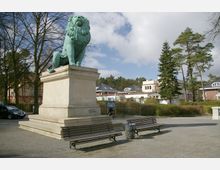 Eine große Löwenskulptur aus grünlicher Bronze auf einem steinernen Sockel in einem offenen Platzbereich, umgeben von Bänken, einem Papierkorb, Bäumen und Wohnhäusern im Hintergrund. Der Himmel ist teils bewölkt, und die Umgebung wirkt gepflegt und ruhig.