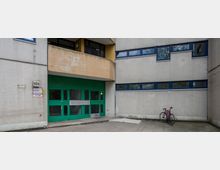 Wohnhausfassade aus Beton mit kleinen gefliesten Quadraten, blau gerahmten Fenstern und einem großen, grünen Eingangstor. Links ein Gebäudeschild mit der Nummer "40h"; rechts steht ein angelehntes Fahrrad neben der Wand.