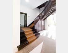 Treppenhaus mit hölzerner Wendeltreppe, schwarzem Geländer und Stufen mit einem beigen Läufer. Links befindet sich eine Türöffnung, rechts eine verglaste Tür, die zu einem Außenbereich führt.