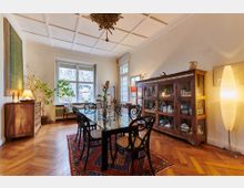 Esszimmer mit holzgetäfeltem Parkettboden, einem Glastisch mit geschwungenen Stühlen und einem orientalischen Teppich. An der Wand steht ein großer, antiker Holzschrank mit Glastüren, daneben eine Stehlampe und einige Pflanzen, während ein großes Fenster den Raum erhellt.