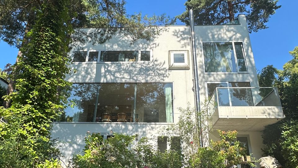 Modernes, weißes Mehrfamilienhaus mit großen Fensterfronten und einem Glasbalkon, umgeben von hohen Bäumen. Im Vordergrund liegt ein üppiger Garten mit Sträuchern und blühenden Pflanzen.
