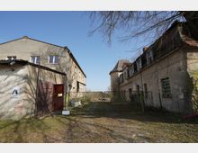 Blick in einen verwitterten Hof zwischen zwei alten, verputzten Gebäuden mit kleinen Fenstern und teils beschädigtem Dach. Ein grünes Metalltor versperrt die Einfahrt; der Boden besteht aus unregelmäßigem Kopfsteinpflaster, darüber ragen kahle Baumäste in den Himmel.