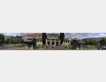 Panoramablick auf den Eingang der historischen U-Bahn-Station Wittenbergplatz in Berlin mit ihrem neoklassizistischen Gebäude, flankiert von zwei kleinen Pavillons. Im Hintergrund sind moderne Gebäude und Bäume zu sehen, während sich im Vordergrund Menschen auf dem Platz aufhalten.