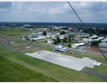 Luftaufnahme eines kleinen Flugplatzes mit Beton-Start- und Parkflächen, mehreren geparkten Kleinflugzeugen und einem Kontrollturm in der Mitte. Umgeben von grasbedeckten Flächen, Straßen und benachbarten Gebäuden in einer ländlichen Umgebung.