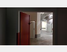 Ein Blick durch eine geöffnete rote Stahltür in einen industriellen Innenraum mit Betonboden, sichtbaren Rohrleitungen an der Decke und mehreren weißen Türen in der Ferne. Die Wände sind schlicht und in hellen Farben gehalten.