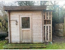 Holzgartenhaus mit Flachdach aus unbehandeltem Holz, ausgestattet mit einer Holztür mit Glaseinsatz und einem kleinen Laternenlicht an der Wand. Rechts befindet sich ein offener Holzlagerbereich, umgeben von einem Garten mit Bäumen und Gras.