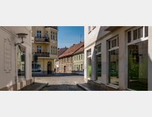 Eine gepflasterte Straßenecke in einem Altstadtviertel mit historischen Gebäuden, darunter ein gelbes Mehrfamilienhaus mit Balkonen im Jugendstil sowie angrenzende Häuser mit roten und grünen Fassaden. Auf der rechten Seite befinden sich moderne Fenster mit Spiegelungen.