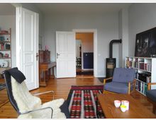 Wohnzimmer mit Holzboden und grauen Wänden, ausgestattet mit einem modernen Sessel mit Fellauflage, einer bunten Teppichdecke, einem Holzklavier, einem kleinen Kaminofen, einem Bücherregal und einem Couchtisch. Im Hintergrund sind geöffnete weiße Doppeltüren zu sehen, die in einen angrenzenden Raum führen.