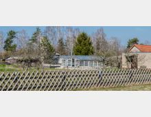 Ein ländlicher Garten mit einem Holzlattenzaun im Vordergrund, einem Gewächshaus aus Glas und Metall in der Mitte und mehreren Gebäuden mit roten Dächern im Hintergrund, umgeben von Bäumen und Wiesen. Im Hintergrund sind Laub- und Nadelbäume zu sehen.