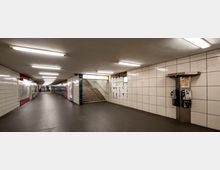 U-Bahn-Unterführung mit weißen, quadratischen Wandfliesen und dunklem Boden. In der Mitte führen Treppen nach oben, an den Wänden hängen Werbetafeln und es stehen zwei öffentliche Telefone.