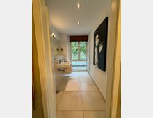 Schmales Badezimmer mit beigen Fliesen, einem Wandwaschbecken links, modernen Spotlights in der Decke und einem großen Fenster mit Holzjalousie am Ende des Raums. Rechts hängt ein schwarz-weißes Gemälde einer Frau.