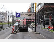 Einfahrt zu einem Q-Park-Parkhaus am Potsdamer Platz in Berlin, mit einem Schild, das freie Plätze anzeigt. Im Hintergrund befinden sich moderne Bürogebäude, eine Straße mit geparkten Fahrzeugen und einige Fahrräder; der Bereich ist von kahlen Bäumen und Gehwegen gesäumt.