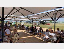 Gruppe von Menschen sitzt unter großen, weißen Sonnenschirmen an Holztischen auf einem Freigelände. Im Hintergrund stehen historische Flugzeuge und Oldtimer auf einer Wiese, umgeben von weiteren Besuchern.