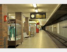 U-Bahn-Station mit einer gewölbten Decke, braunen Kachelsäulen und einem Bahnsteig mit Sitzgelegenheiten. Zu sehen sind ein Fahrplan, eine Uhr, Geschäfte sowie ein Schaukasten mit Kleidung; auf der rechten Seite verlaufen die Bahngleise.