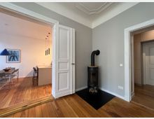 Wohnraum mit Holzboden und hellgrauen Wänden. Im Vordergrund steht ein freistehender schwarzer Kaminofen auf einer schwarzen Bodenplatte, links führt eine geöffnete zweiflügelige Tür in den Essbereich mit einem großen Esstisch, einer Hängelampe und einer modernen Küche im Hintergrund.