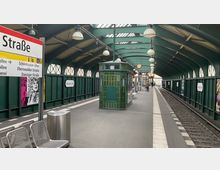 Bahnhof "Eberswalder Straße" in Berlin mit grün gestrichener Überdachung und gewölbtem Dach, flankiert von hohen Fenstern. Im Vordergrund Sitzbänke, ein Abfalleimer und ein grün gefliestes Betriebsgebäude; im Hintergrund Gleise, Laternen und ein Uhrenschild.