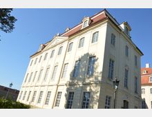 Helles Barockgebäude mit mehreren großen, symmetrisch angeordneten Fenstern und einem roten Mansarddach, umgeben von einem klaren blauen Himmel. Im Vordergrund sind Teile eines Laternenpfahls und Schatten von Bäumen sichtbar.