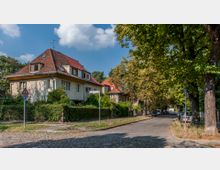 Wohnviertel mit freistehenden Einfamilienhäusern, roten Ziegeldächern und umgebenden Gärten entlang einer gepflasterten Straße. Rechts begrenzt eine Reihe von Bäumen die Fahrbahn, während ein Straßenschild und geparkte Autos sichtbar sind.