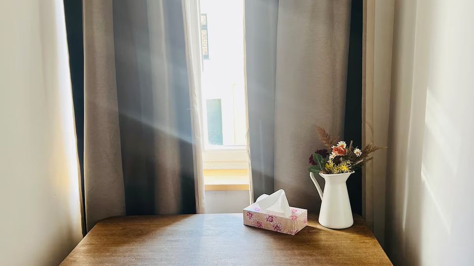 Kleine Sitzecke in einem Innenraum mit einem einfachen Holztisch vor einem hohen Fenster, das von dunklen Vorhängen eingerahmt ist. Auf dem Tisch stehen eine Taschentuchbox und eine weiße Kanne mit Trockenblumen, durch das Fenster fällt Sonnenlicht ein.