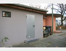 Flaches Gebäude mit grauer, glatter Fassade, einer Metalltür und einem kleinen, rechteckigen Fenster. Im Vordergrund stehen eine Mülltonne und ein Einkaufswagen, daneben ein abgerundetes, rosafarbenes Gebäude mit Vordach sowie eine Pflasterfläche mit Bäumen im Hintergrund.