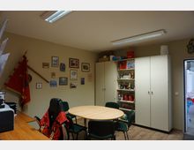 Ein kleiner Besprechungsraum mit cremefarbenen Wänden, einem runden Holztisch und mehreren Stühlen. An der Wand hängen Bilder und Vereinsutensilien, daneben stehen Aktenschränke und eine Vitrine mit Pokalen und Büchern; durch eine offene Tür ist ein angrenzender Raum erkennbar.