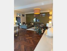 Wohnzimmer mit modernem Interieur, dunklem Fischgrätenparkett und einer großen, grauen Eckcouch. Die Wände sind olivgrün gestrichen, an denen runde Spiegel angebracht sind, und das Zimmer ist mit Pflanzen, Pendelleuchten und dekorativen Elementen ausgestattet.