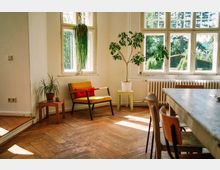 Heller Raum mit Holzboden und großen Fenstern, durch die grünes Außenlicht hereinfällt. Im Vordergrund stehen ein gelber Sessel mit rotem Kissen, ein kleiner Tisch mit einer Topfpflanze, sowie ein Esstisch mit Stühlen; weitere Pflanzen dekorieren den Raum.