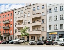 Straßenansicht mehrerer mehrstöckiger Wohnhäuser mit hellen Fassaden und gestaffelten Balkonen, teils mit Blumenkästen. Im Vordergrund stehen geparkte Autos, ein kleiner Straßenbaum und eine Laterne; im Erdgeschoss sind Ladenfronten mit Schaufenstern zu sehen.