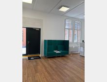 Ein heller Raum mit Holzfußboden, einer schwarzen Eingangstür links und großen Fenstern mit weißen Rahmen, die Tageslicht hereinlassen. Rechts steht eine Sitzbank mit grünen Polstern und integriertem kleinen Tisch, daneben ein weißes Regal.