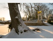 Winterliche Parklandschaft mit verschneitem Baum im Vordergrund, einer steinernen Plattform mit Geländer im Hintergrund und einer vergoldeten Stierstatue auf einem kleinen Tempel mit Säulen. Die Szene ist bei Sonnenaufgang oder Sonnenuntergang beleuchtet.