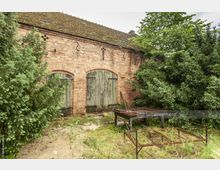 Alte Backsteinfassade mit drei großen, bogenförmigen Holztoren, umgeben von dichter Vegetation. Im Vordergrund befinden sich verrostete Metallgestelle und ein verwitterter Holztisch auf einer bewachsenen Fläche.
