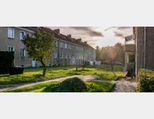 Mehrfamilienhäuser mit grauen Fassaden, roten Ziegeldächern und kleinen Fenstern umgeben eine grüne Rasenfläche mit Wäscheleinen und vereinzelten Bäumen. Im Vordergrund führen Gehwege durch die Wiese, im Hintergrund sind geparkte Autos zu sehen, während die Sonne tief steht und die Szene beleuchtet.