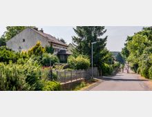 Straßenszene in einem Wohngebiet mit einem weißen, älteren Haus auf der linken Seite, umgeben von Gärten und dichten Bäumen. Eine schmale asphaltierte Straße mit Straßenlaternen führt durch die grüne Umgebung, in der Ferne sind zwei Personen zu Fuß unterwegs.