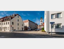 Straßenszene in einer kleinen Stadt mit hellen Gebäuden im traditionellen Stil und roten Ziegeldächern, im Hintergrund ist ein Kirchturm sichtbar. Im Vordergrund befinden sich ein Parkplatz mit Autos, Gehwege und Straßenschilder.
