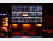 Regal mit beleuchteten Glasböden, auf denen verschiedene Getränkeflaschen stehen; im Hintergrund eine Struktur aus schimmernden LEDs. Unterhalb befindet sich eine Bartheke mit Gläsern und weiteren Barutensilien, rechts ein beleuchteter Bereich mit einer weißen Box.