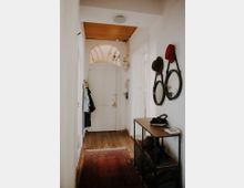 Schmaler Eingangsflur mit Holzboden und weiß gestrichenen Wänden. Am Ende des Flurs befindet sich eine weiße Tür mit einem zugbogenförmigen Glaselement, auf der rechten Seite ein niedriger Schuhschrank mit Ablage und runden Wandspiegeln, links eine Garderobe mit hängenden Jacken. Ein roter Teppich liegt auf dem Boden.