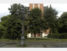 Historischer Platz mit Kopfsteinpflaster, einer verzierten schwarzen Laterne im Vordergrund und einer roten Backsteinkirche, teilweise von dichtem grünen Laub verdeckt, im Hintergrund.