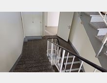 Ansicht eines Treppenhauses mit grobkörnigem Terrazzo-Bodenbelag und weiß gestrichenen Wänden. Offene Tür zu einem angrenzenden Raum sichtbar, ein Metallgeländer mit schwarzen Handläufen führt entlang der Treppe.