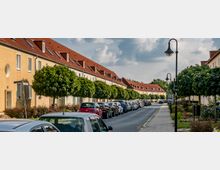 Reihenhaussiedlung mit gelb verputzten Gebäuden und roten Satteldächern entlang einer Straße. Autos parken auf beiden Seiten der Fahrbahn, während regelmäßig begrünte Bäume und Straßenlaternen die Gehwege säumen.