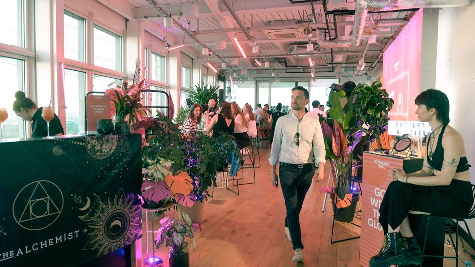 Bright modern event space with large floor-to-ceiling windows, wooden floors, and an exposed industrial ceiling with ducts and hanging lights. Plants and vendor-style counters line the room, with bar stools and a pink-lit projection wall at the far end.