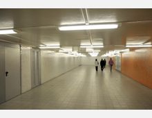 Ein beleuchteter U-Bahn-Tunnel mit weißen Decken- und Wandfliesen sowie orangefarbenen Akzenten auf einer Wand. Vier Personen laufen in der Mitte des Tunnels auf den Ausgang zu.