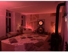 Ein gemütlich eingerichteter Raum mit gedimmtem, rötlichem Licht. Der Raum enthält weiche Teppiche und Kissen auf dem Boden, eine Sitzgelegenheit, ein großes Gong-Instrument, sowie einen Kaminofen an der rechten Wand. Die Wände sind teilweise mit einer strukturierten, künstlerischen Gestaltung versehen.