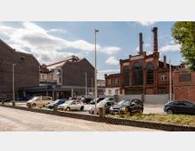 Ein Parkplatz mit mehreren abgestellten Autos, umgeben von Gebäuden im Hintergrund, darunter ein rotes Backsteingebäude mit großen, bogenförmigen Fenstern und Schornsteinen sowie ein graues Gebäude mit einem ALDI-Markt. Der Himmel ist teils bewölkt, und ein Baum ist am rechten Bildrand sichtbar.