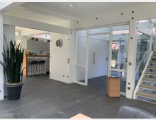 Helles, modernes Wohnzimmer mit grauem Steinboden, einer Glas-Schiebetür und einer angrenzenden offenen Küche mit Barhockern. Rechts führt eine Treppe mit Teppichstufen nach oben, links steht eine große Zimmerpflanze in einem schwarzen Topf.