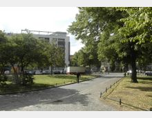 Städtischer Platz mit Kopfsteinpflasterweg, umgeben von Grünflächen und Bäumen. Im Hintergrund sind moderne Gebäude, parkende Autos und ein mobiler Verkaufsstand zu sehen.