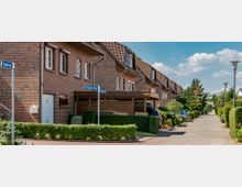 Reihenhäuser aus rotem Klinker mit Satteldächern entlang einer gepflasterten Wohnstraße, gesäumt von Hecken, Gärten und geparkten Autos; Straßenschilder weisen auf den „Knupperweg“ hin.