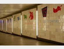 Unterführung mit hellen Fliesenwänden, auf denen stilisierte, farbige Illustrationen und die Namen berühmter Komponisten wie Giuseppe Verdi und Igor Strawinsky abgebildet sind; vertikale Leuchtstreifen trennen die einzelnen Abschnitte.